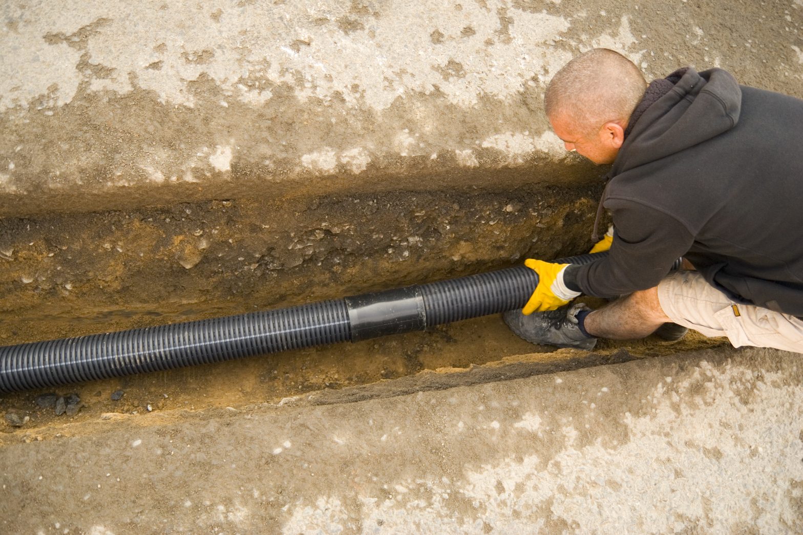 man installing an exterior french drain pipe in a drainage ditch before the stone is covered